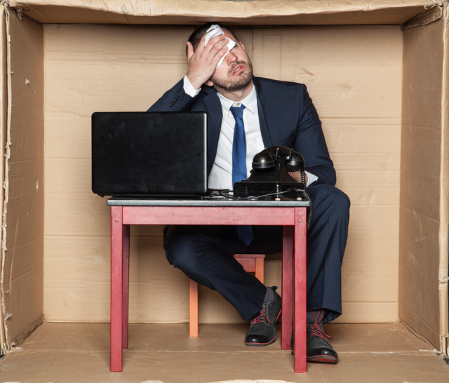 Homme d'affaires assis à un bureau dans une boîte, espace restreint, stress au travail et augmentation de la charge de travail, ordinateur et téléphone, homme s'essuyant le front.