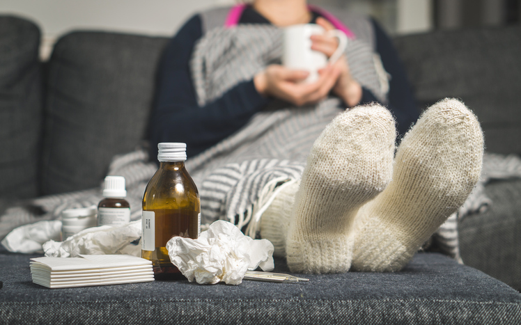 une femme malade est allongée dans son lit, avec des chaussettes chaudes, des pilules et des sirops à côté d'elle