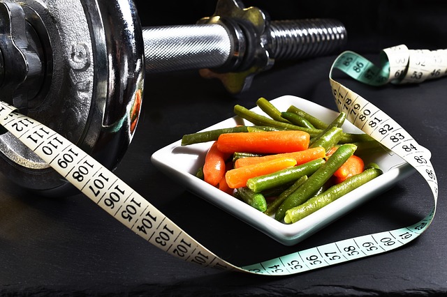 Un haltère chromé placé à côté d'une assiette carrée. Sur l'assiette se trouve un légume, à côté duquel se trouve un mètre de tailleur.