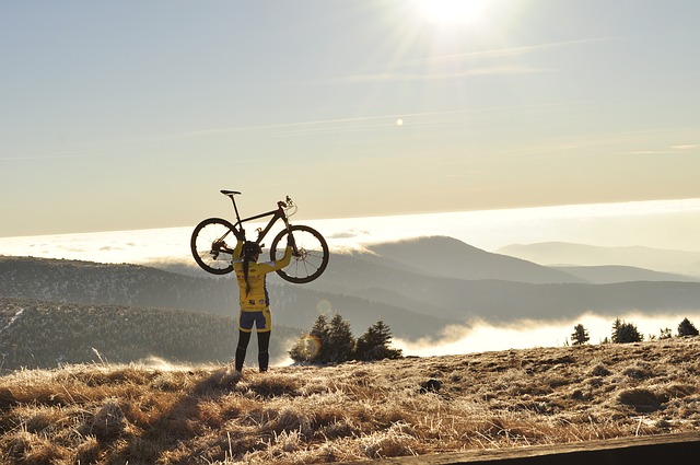 Une cycliste soulève son vélo au-dessus de sa tête, avec en arrière-plan des montagnes dans le brouillard.