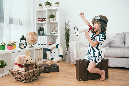 Une petite fille vêtue d'un manteau en peau de mouton, assise sur une valise dans le salon et tenant un mégaphone.