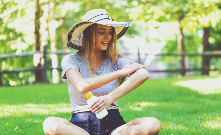 Une jeune femme se met de la crème solaire, assise dans un pré, en portant un chapeau pour se protéger la tête.