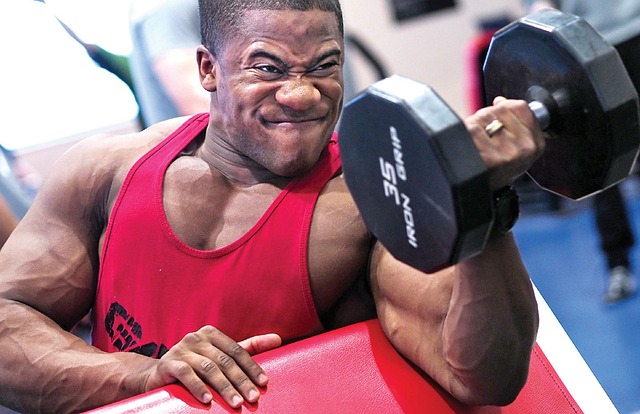 Homme en débardeur rouge faisant des biceps avec une barre d'haltères à un bras.