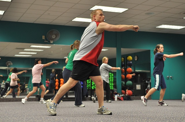 Cinq personnes font de la musculation dans la salle de sport. Un homme et quatre femmes. Ils sont habillés de manière athlétique, dos à un grand miroir. Le mur est vert et la moquette au sol est grise.