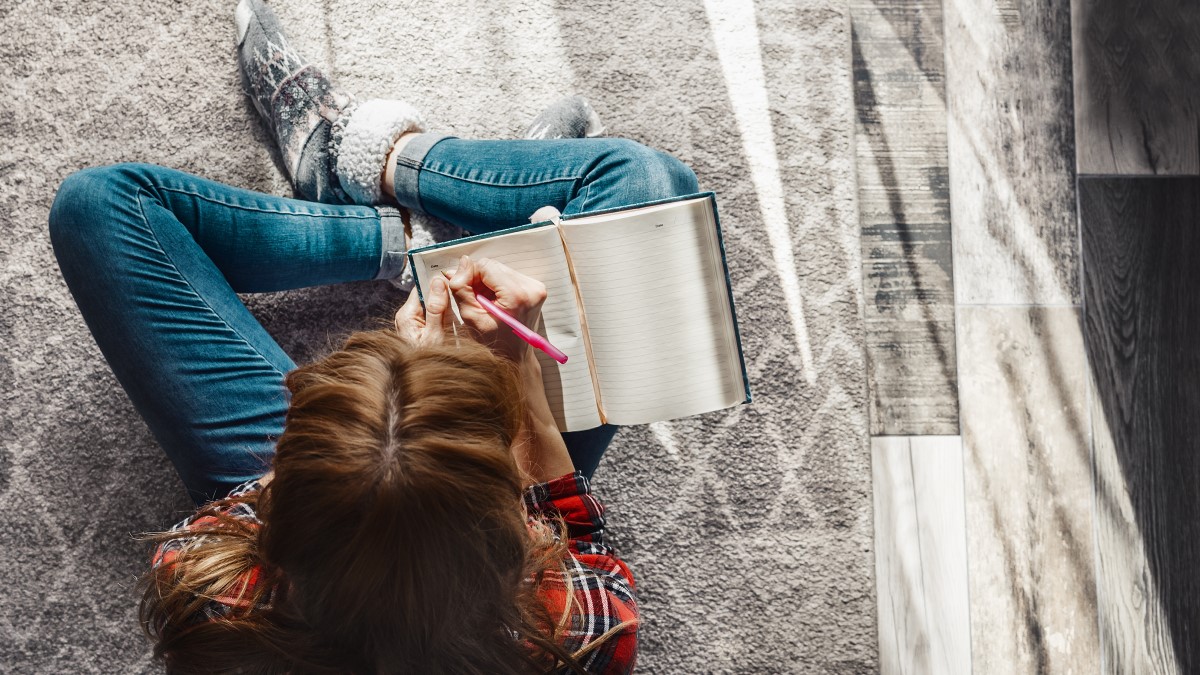 Une femme est assise sur un tapis, tient un carnet et note des détails, consignant les maux de tête dans le journal d'une migraineuse.