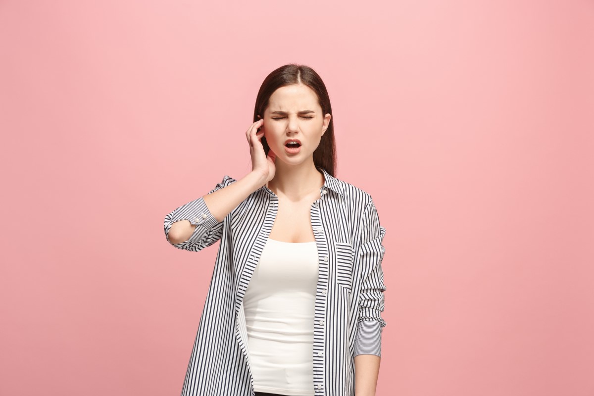 Une femme a mal à l'oreille et la tient. Elle est debout. Fond rose.