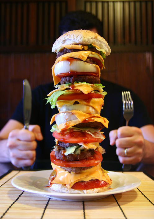 Un homme est assis à une table avec un grand hamburger dans son assiette, une mégapuissance, une voiture et une voiture.