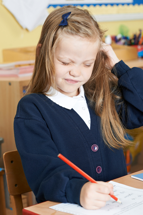 Une petite fille assise à l'école avec des démangeaisons à la tête, probablement des poux dans les cheveux, des cheveux blonds, assise et écrivant.