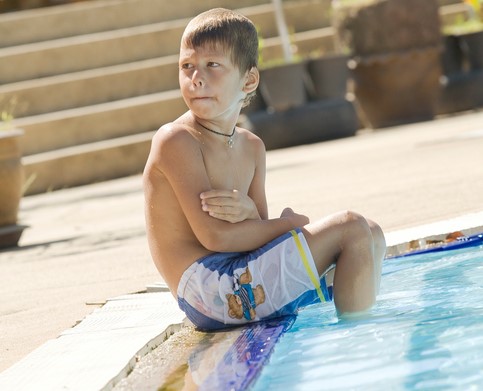 Le garçon est assis au bord de la piscine, les pieds dans l'eau. Il a froid.